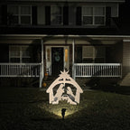 Crèche de Noël illuminée devant une maison en brique, avec personnages en silhouette blanche, étoile au sommet, ambiance festive nocturne.