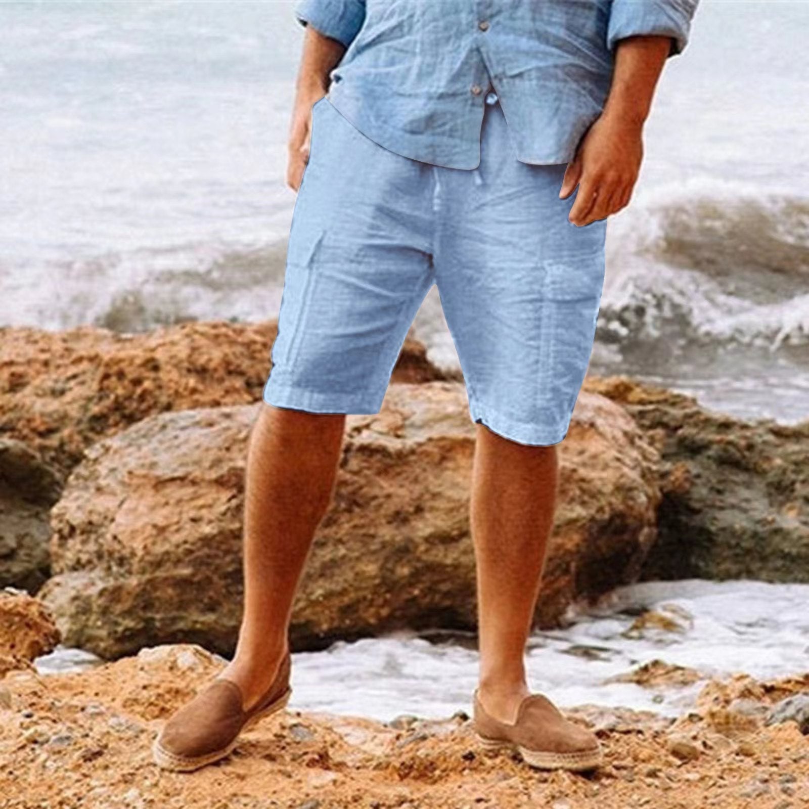 Homme en tenue estivale décontractée, chemise et short en lin bleu, debout sur des rochers près de la mer, style plage, mode homme été.