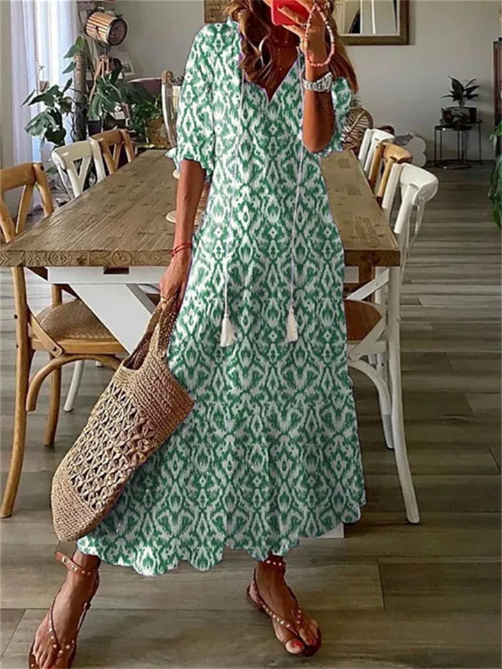 Robe longue bohème verte à motifs, femme tenant un sac en osier, sandales, intérieur moderne avec table en bois et chaises, ambiance estivale.