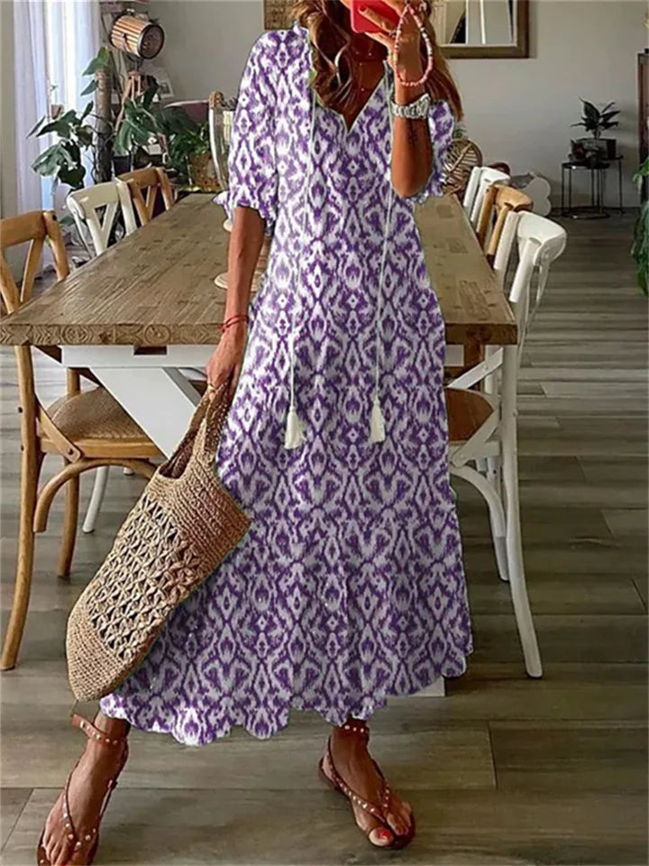 Robe longue bohème violette à motifs, femme tenant un sac en osier, sandales, intérieur moderne avec table en bois et chaises.