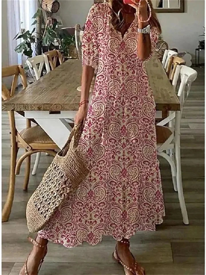 Robe longue bohème à motifs floraux rouges et blancs, femme tenant un sac en osier, sandales, intérieur moderne avec table en bois et chaises.