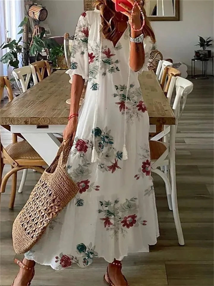 Robe longue bohème blanche à motifs floraux rouges et verts, femme tenant un sac en osier, intérieur moderne avec table en bois et chaises.