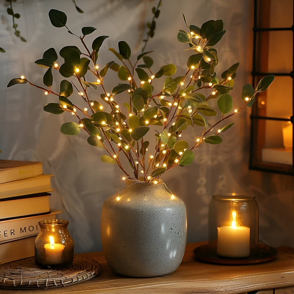 Vase en céramique grise avec branches décoratives illuminées par des guirlandes LED, entouré de bougies allumées sur une table en bois.