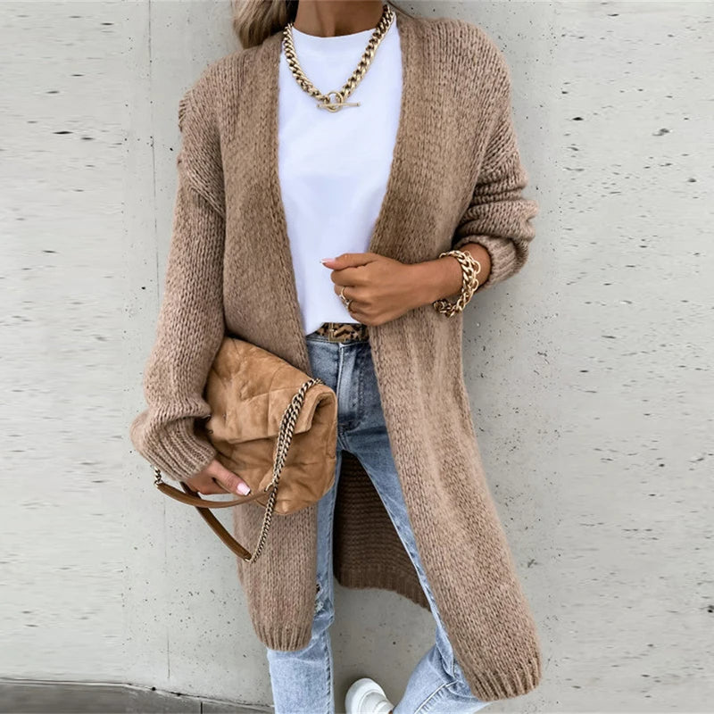 Femme portant un long cardigan beige, t-shirt blanc, jeans décontracté, sac à main en daim marron, accessoires dorés, posant contre un mur gris.