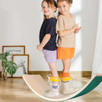 Deux enfants souriants en t-shirts et shorts colorés, debout sur une planche d'équilibre en bois dans un salon lumineux. Jouets éducatifs, jeu intérieur.
