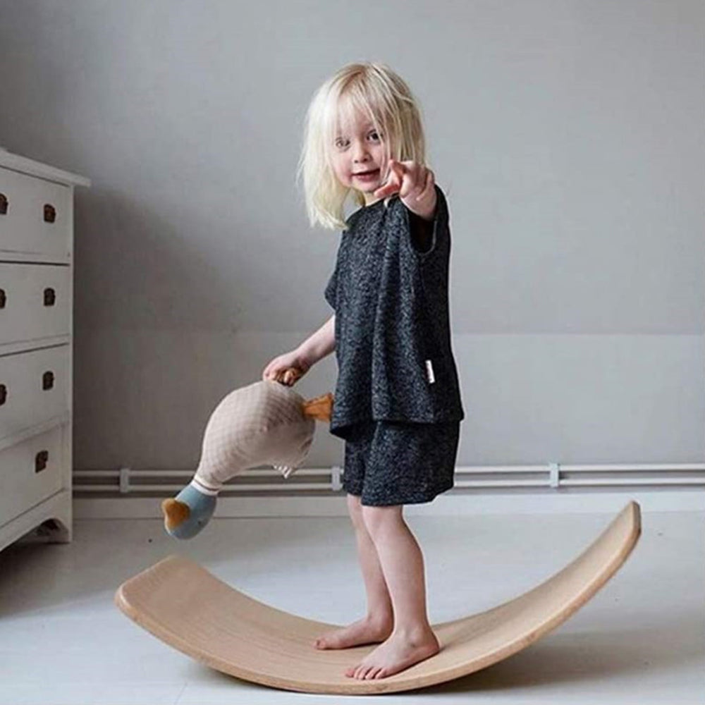 Enfant blond jouant sur une planche d'équilibre en bois, tenant une peluche. Chambre d'enfant moderne, mobilier minimaliste.
