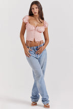 Jeune femme portant un haut rose à manches bouffantes et un jean bleu clair, posant debout sur fond blanc. Mode féminine, style décontracté.