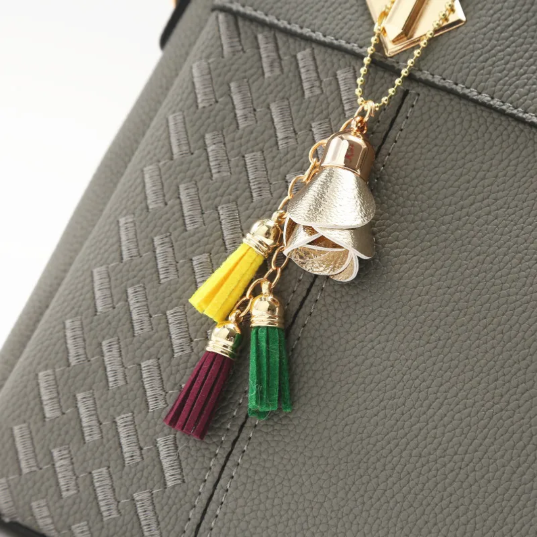Pendentif floral doré sur sac à main gris texturé, avec pompons colorés jaune, vert et bordeaux. Accessoire mode élégant et tendance.