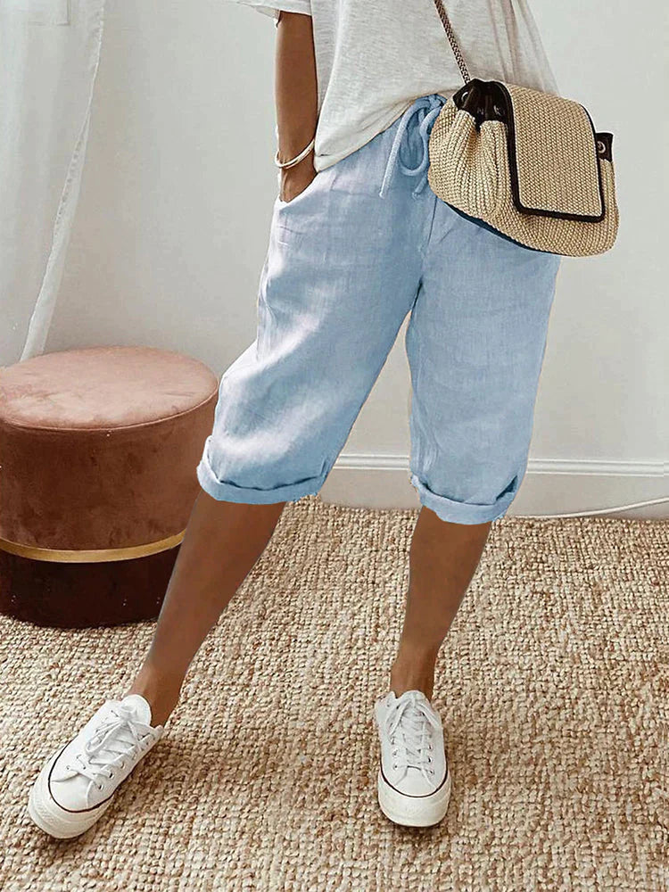 Femme en short bleu clair et t-shirt blanc, portant un sac en osier, baskets blanches, posant sur un tapis beige. Mode décontractée estivale.