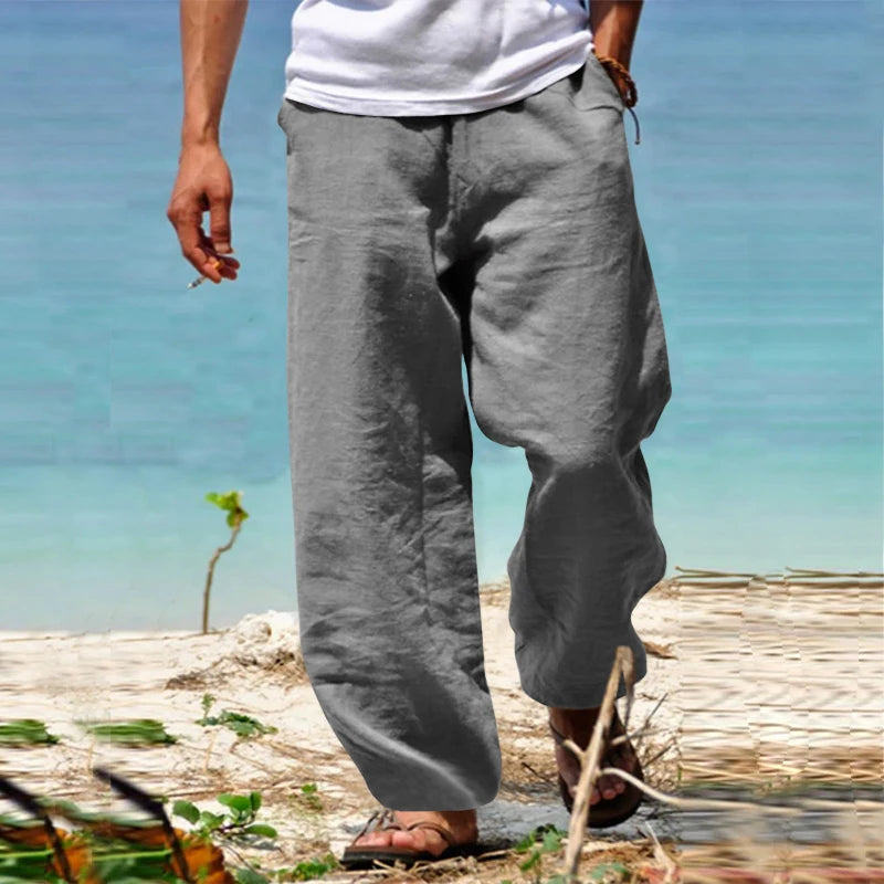 Homme portant un pantalon en lin gris, marchant sur une plage ensoleillée. Mode estivale décontractée, style plage, confort et élégance.
