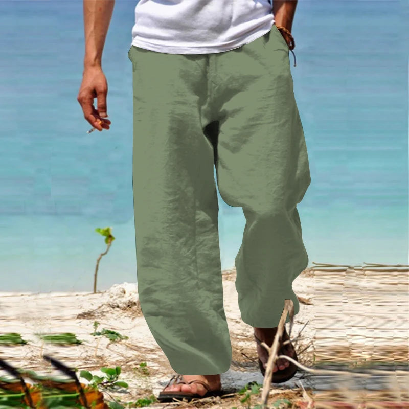 Homme portant un pantalon en lin vert sur une plage, pieds nus, avec une mer turquoise en arrière-plan. Mode estivale décontractée, style plage.