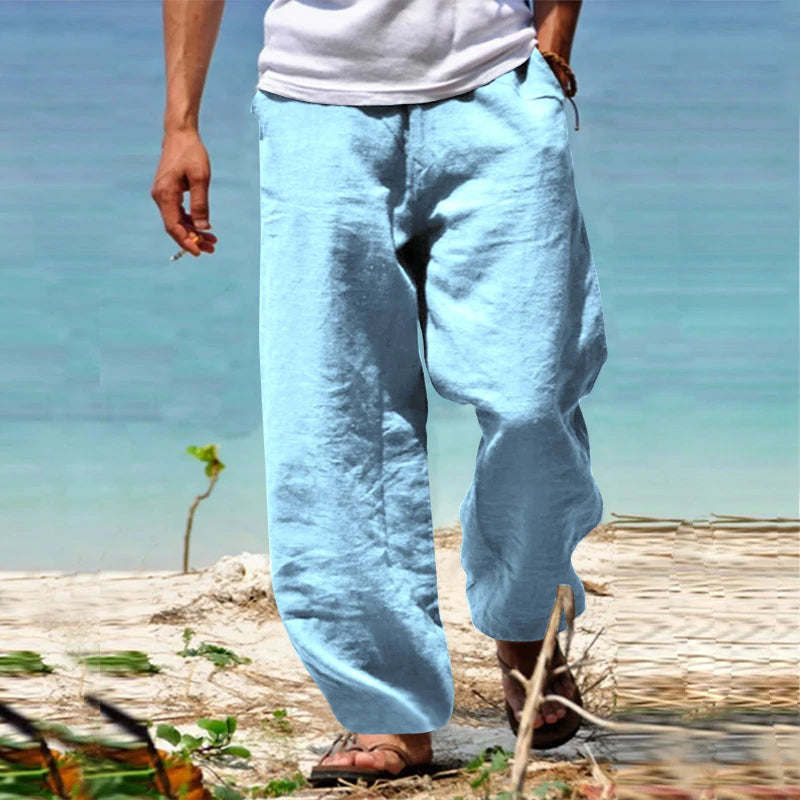 Homme portant un pantalon en lin bleu clair sur une plage, pieds nus, avec vue sur la mer. Mode estivale décontractée, tenue de plage élégante.