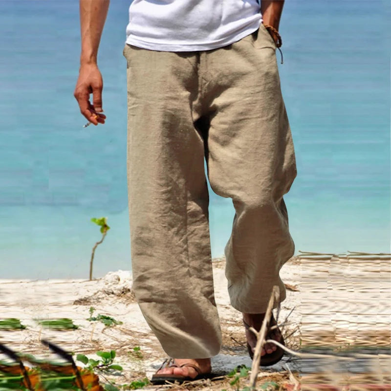 Homme portant un pantalon en lin beige sur une plage, avec un t-shirt blanc et des sandales. Mode estivale décontractée, bord de mer.