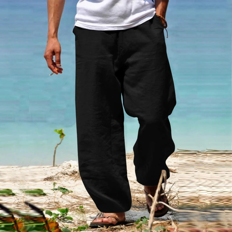 Homme en pantalon noir ample sur plage, pieds nus, chemise blanche, mer turquoise en arrière-plan. Mode décontractée, vacances, été.