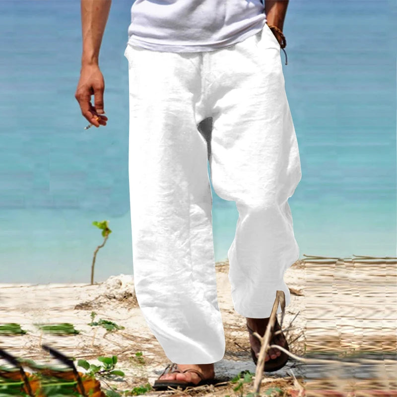 Homme en pantalon blanc en lin marchant sur une plage, portant un t-shirt blanc et des sandales, avec la mer turquoise en arrière-plan.