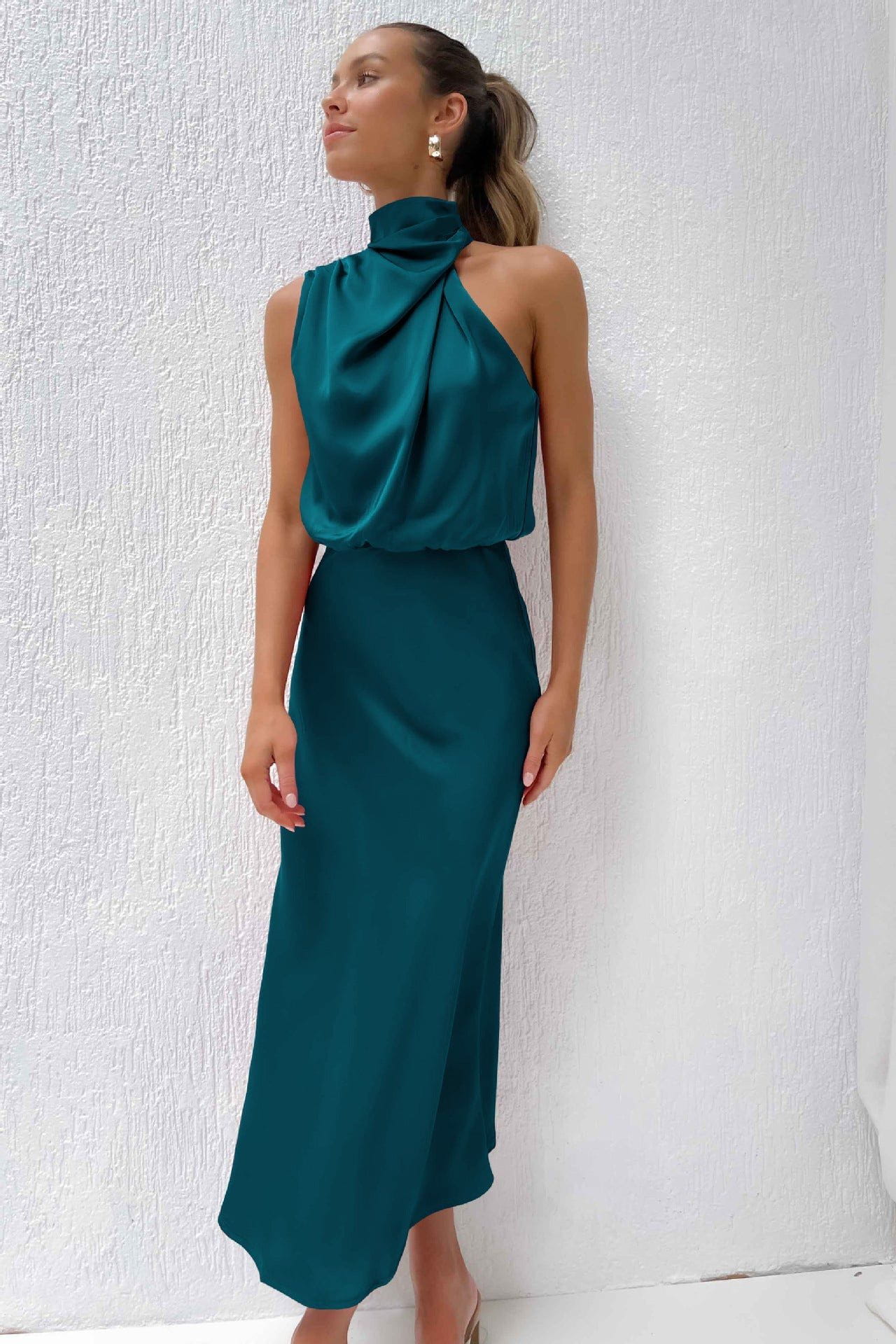 Robe longue en satin vert émeraude, col haut drapé, portée par une femme élégante, fond blanc texturé. Mode féminine chic et tendance.