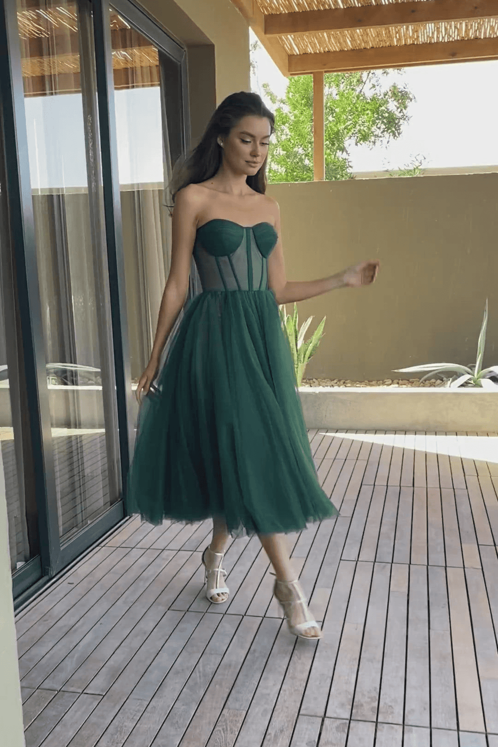 Femme en robe verte élégante marchant sur une terrasse en bois, devant une grande baie vitrée, par une journée ensoleillée.