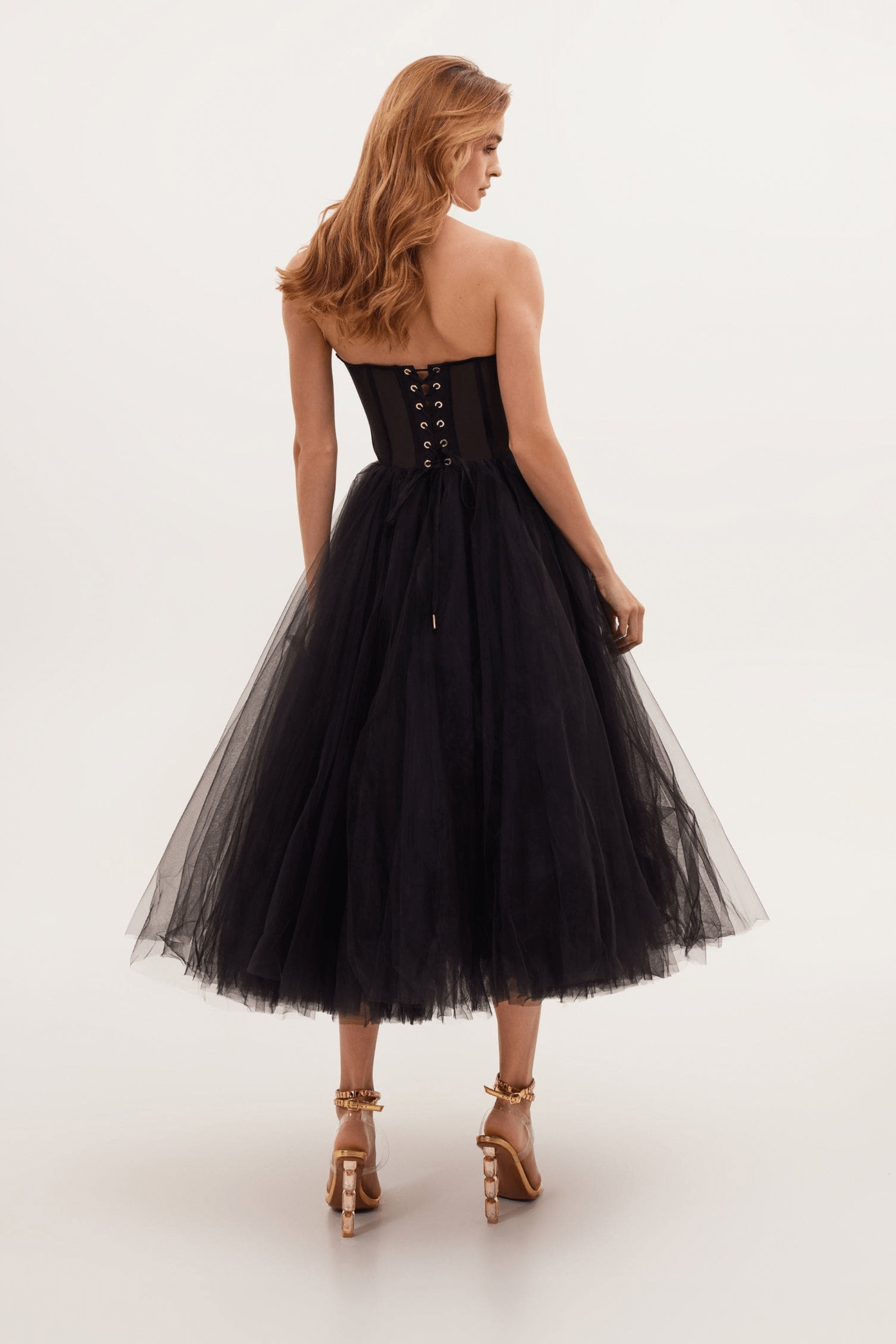 Femme en robe noire bustier élégante, jupe en tulle volumineuse, vue de dos, cheveux longs ondulés, sandales à talons dorés.