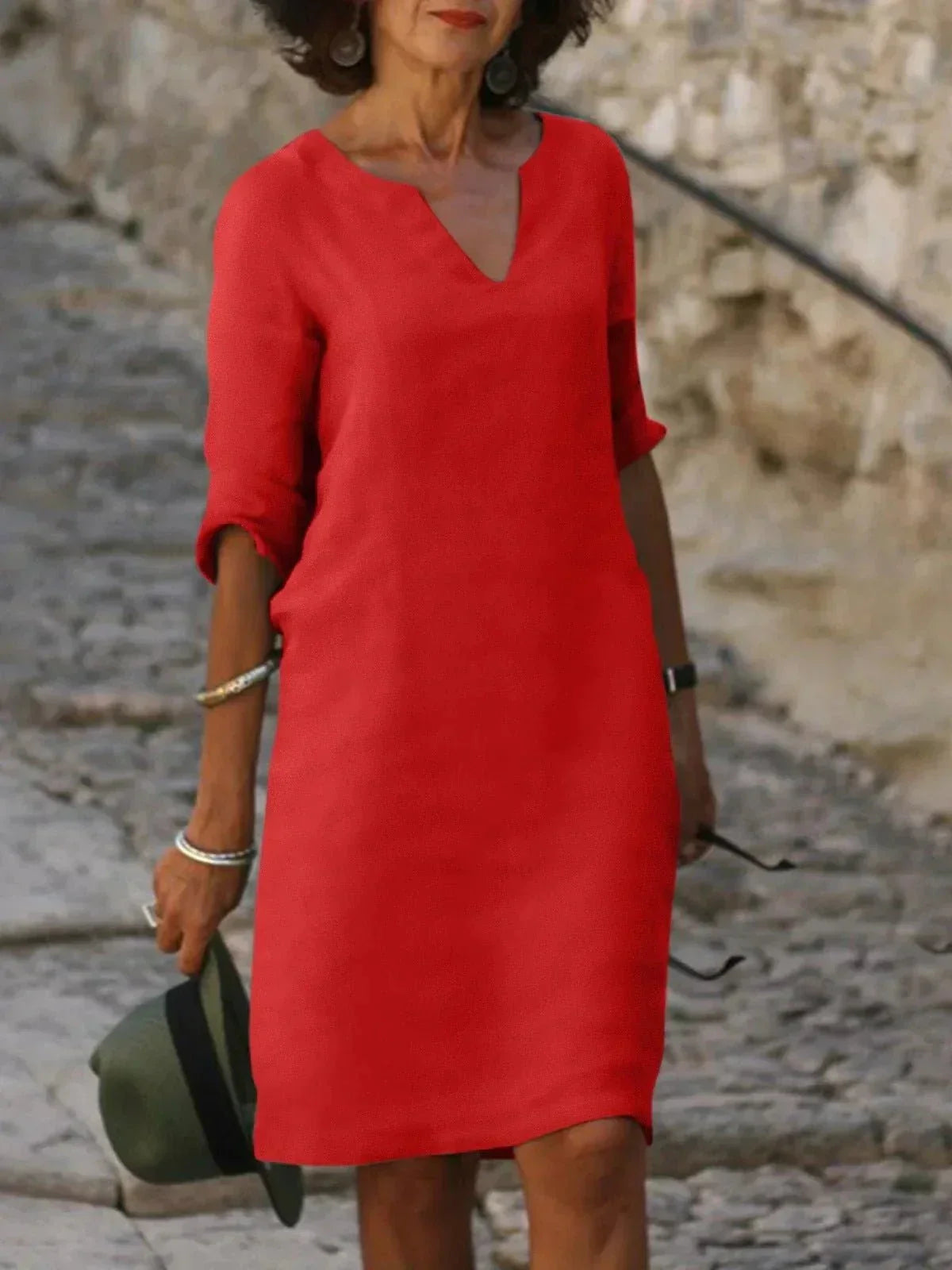 Femme portant une robe rouge élégante, manches retroussées, tenant un chapeau vert. Mode féminine chic, style décontracté, rue pavée en arrière-plan.