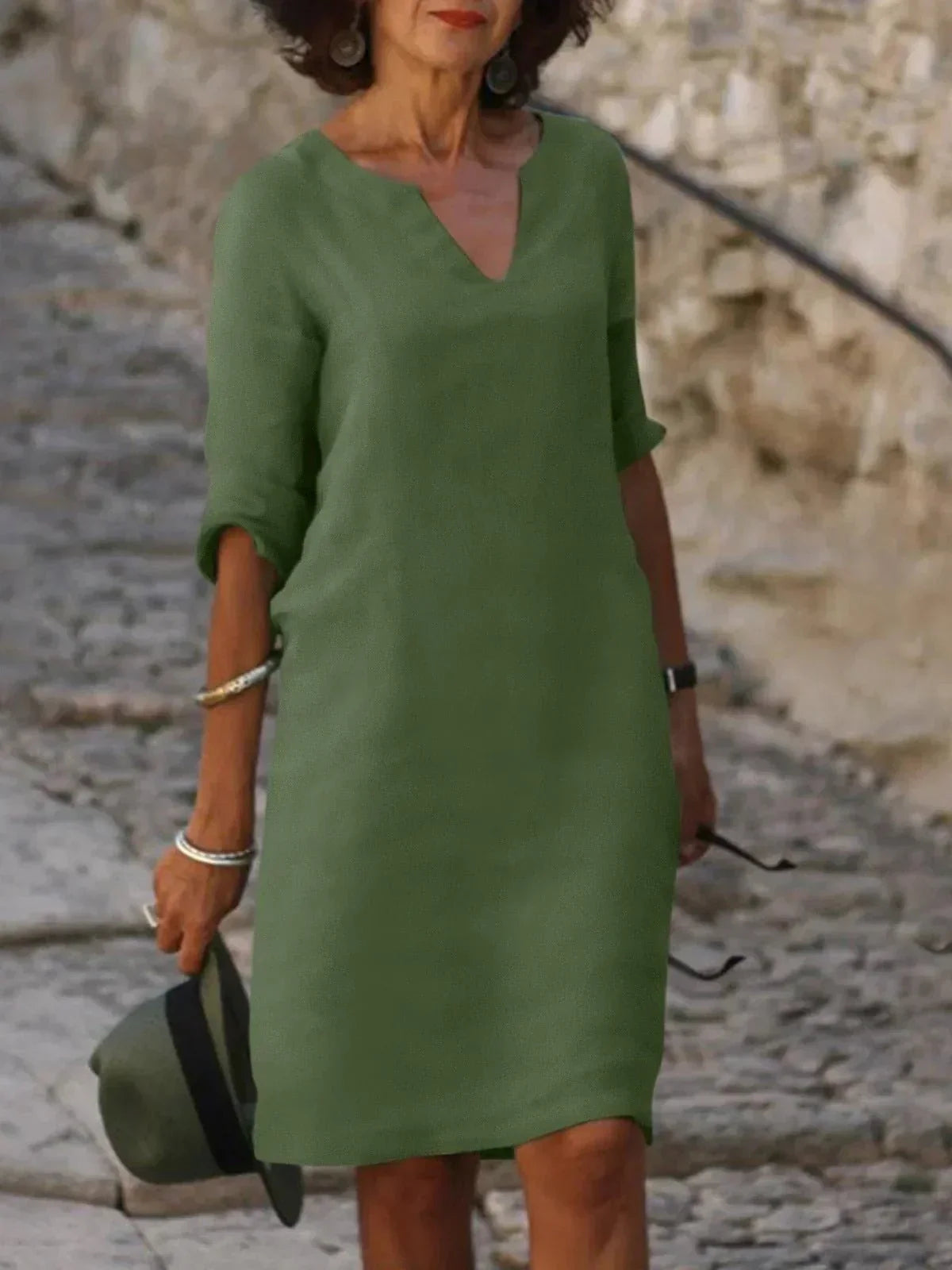 Femme portant une robe verte élégante, manches trois-quarts, tenant un chapeau. Mode féminine chic, style décontracté, promenade sur chemin pavé.