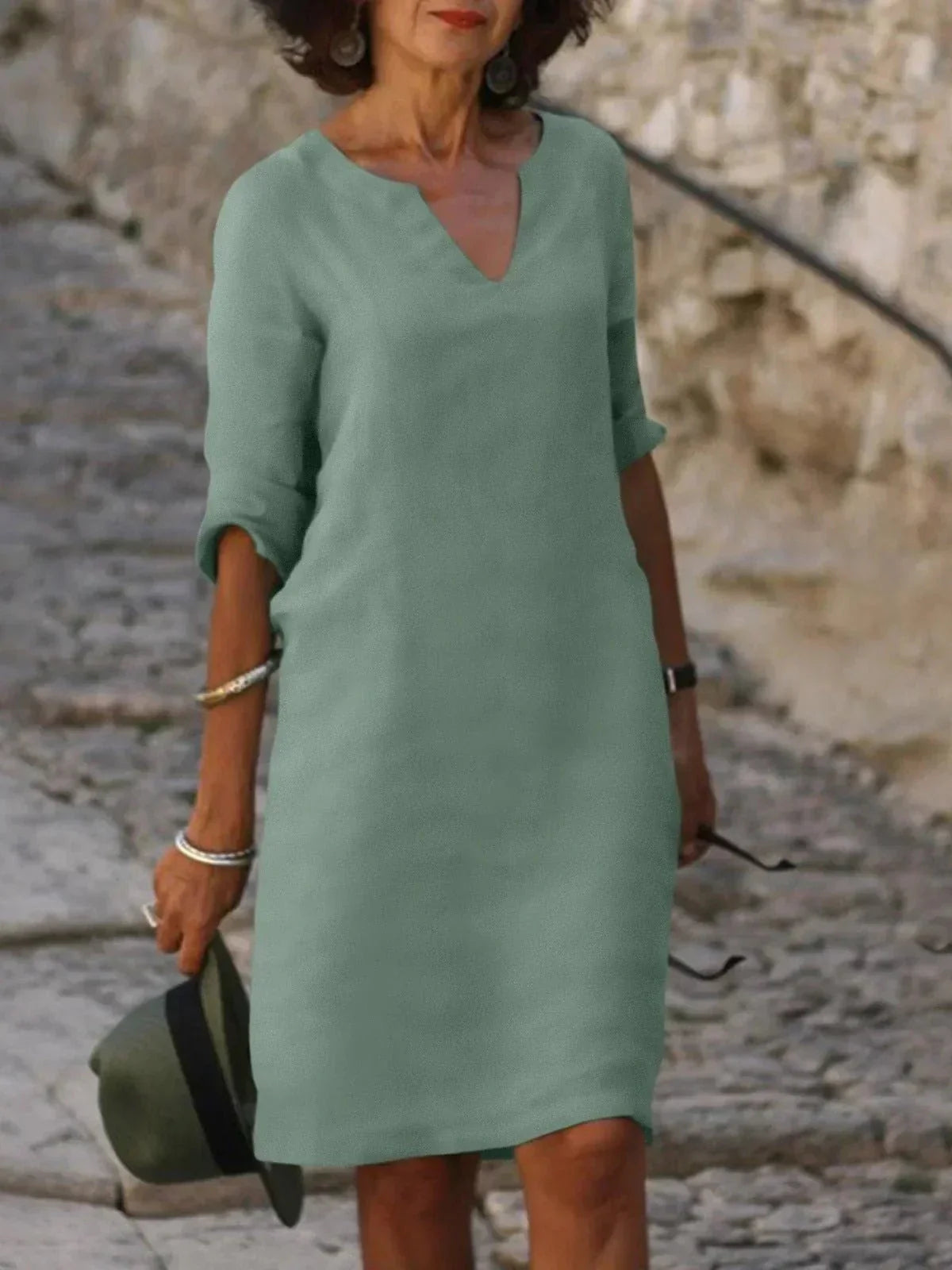 Femme portant une robe verte élégante, manches trois-quarts, tenant un chapeau et des lunettes de soleil, marchant sur un chemin pavé. Mode féminine chic.