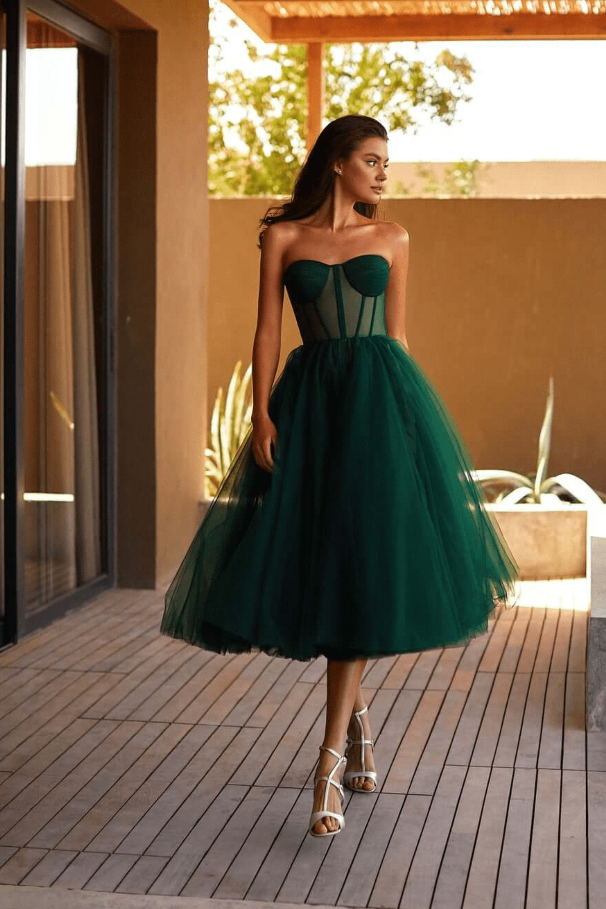 Femme en robe bustier verte élégante, jupe en tulle, marchant sur une terrasse en bois, style chic et moderne, sandales blanches, journée ensoleillée.