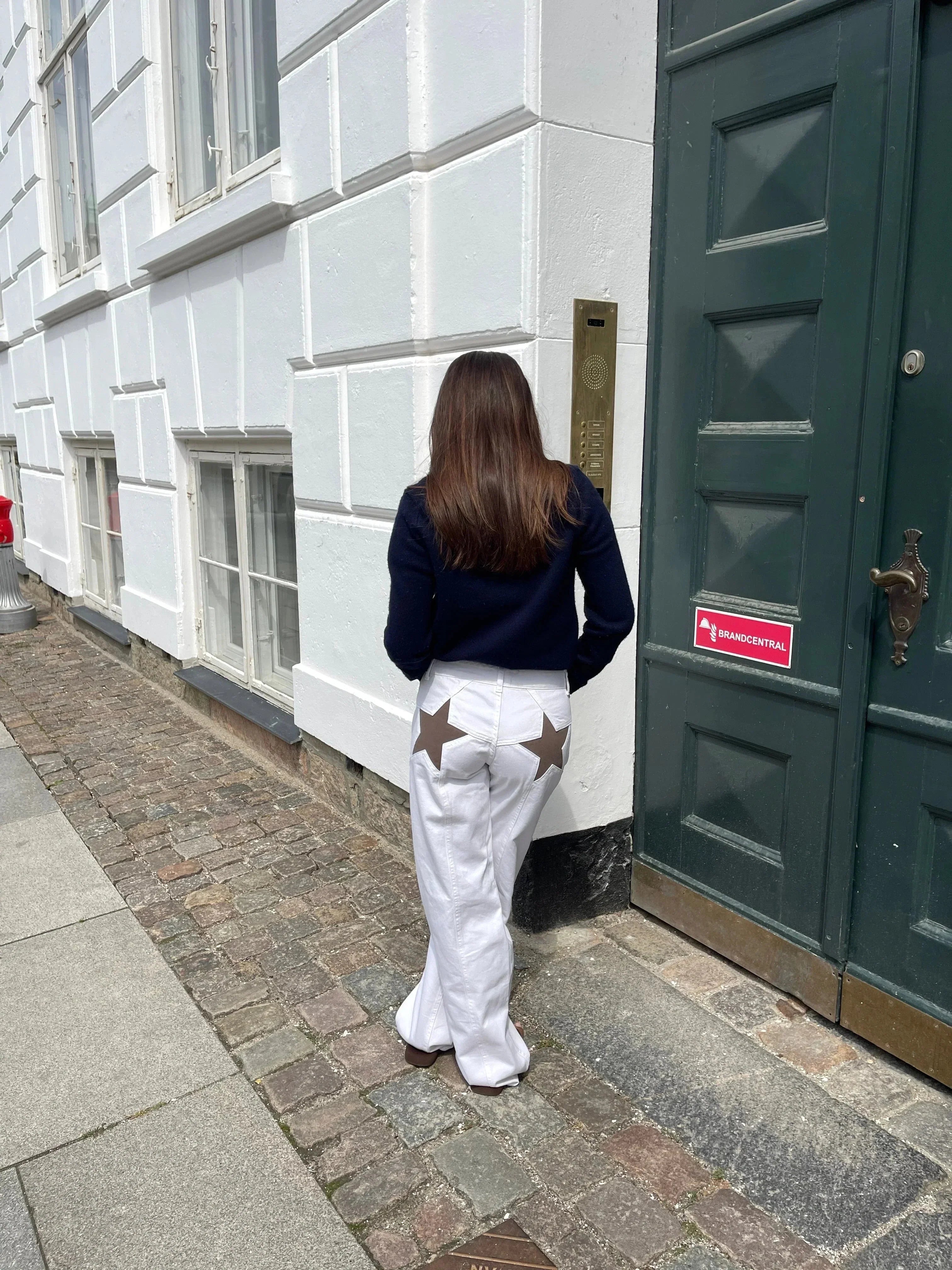 Femme de dos portant un pull noir et un pantalon blanc avec étoiles marron, debout près d'une porte verte et d'un mur blanc en ville.