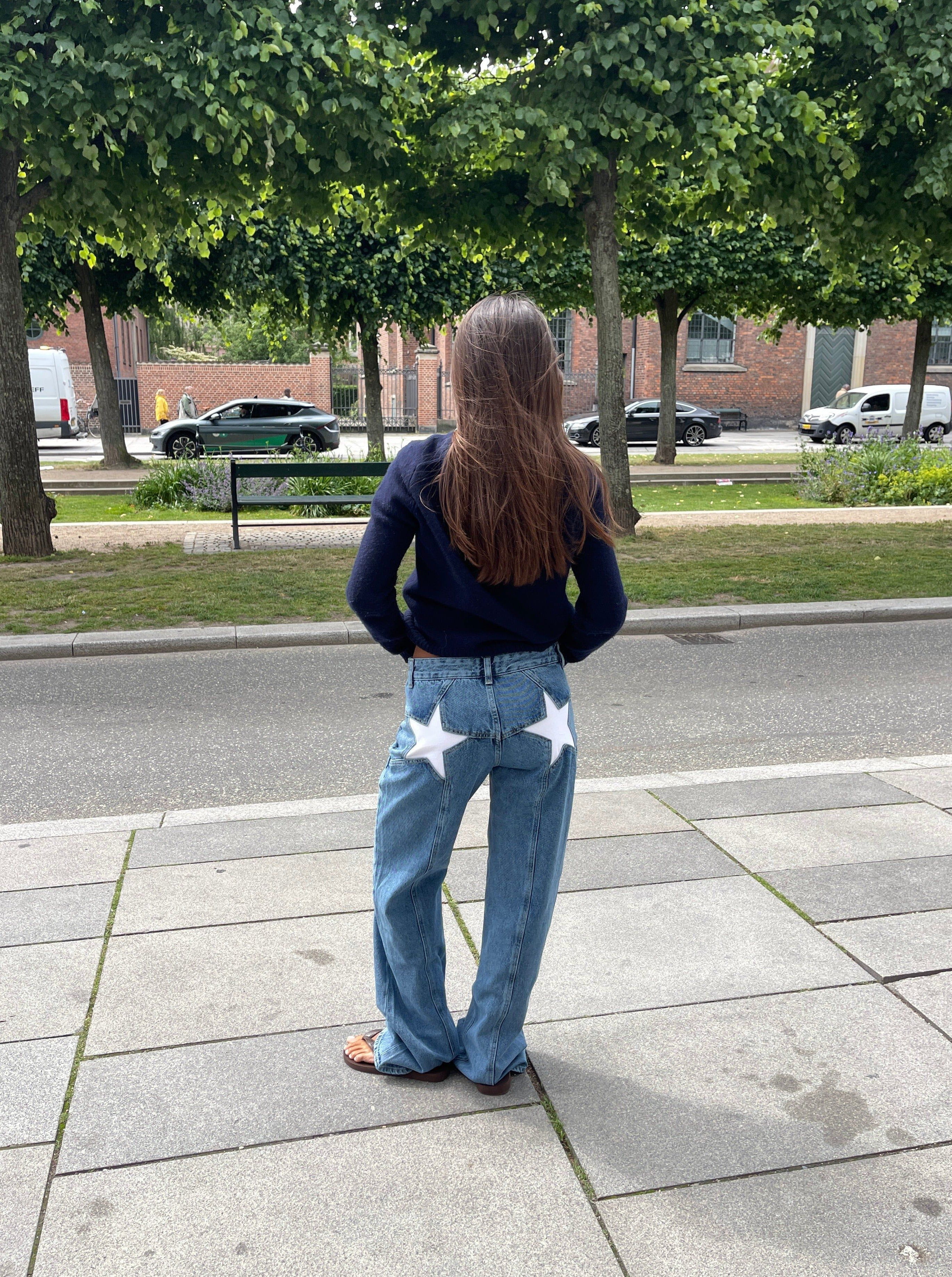 Femme de dos portant un jean bleu avec étoiles blanches, debout sur un trottoir urbain, arbres et voitures en arrière-plan. Mode décontractée.