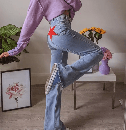 Femme en jean évasé bleu avec étoile rouge, pull violet, posant dans un intérieur moderne avec plantes et fleurs. Mode tendance, style décontracté.