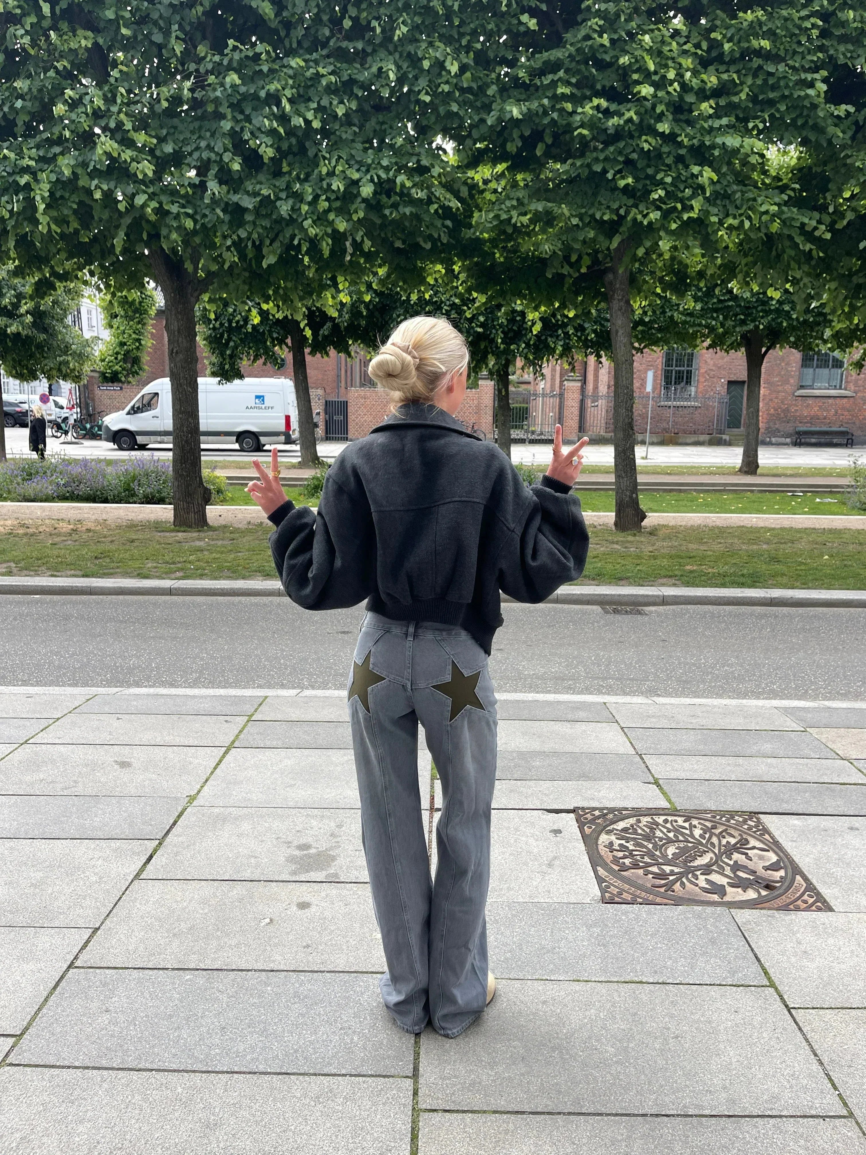 Femme blonde de dos portant une veste noire et un jean gris avec étoiles, posant sur un trottoir devant des arbres et bâtiments urbains.