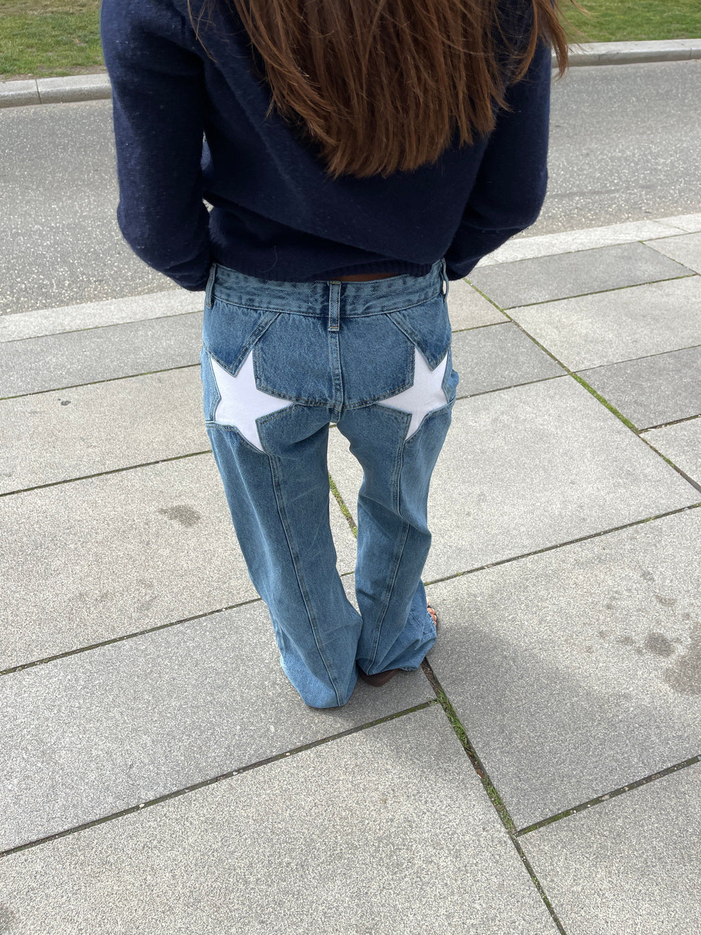 Jeune femme portant un jean bleu avec des étoiles blanches à l'arrière, debout sur un trottoir. Mode urbaine, style décontracté, tendance.