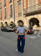 Femme en jean étoilé rouge et t-shirt gris, debout devant des voitures de sport Porsche, rue pavée, architecture parisienne en arrière-plan.