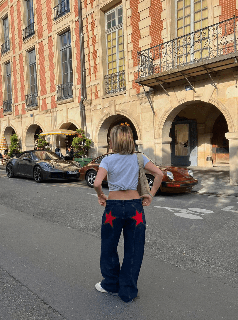 Femme en jean étoilé rouge et t-shirt gris, debout devant des voitures de sport Porsche, rue pavée, architecture parisienne en arrière-plan.