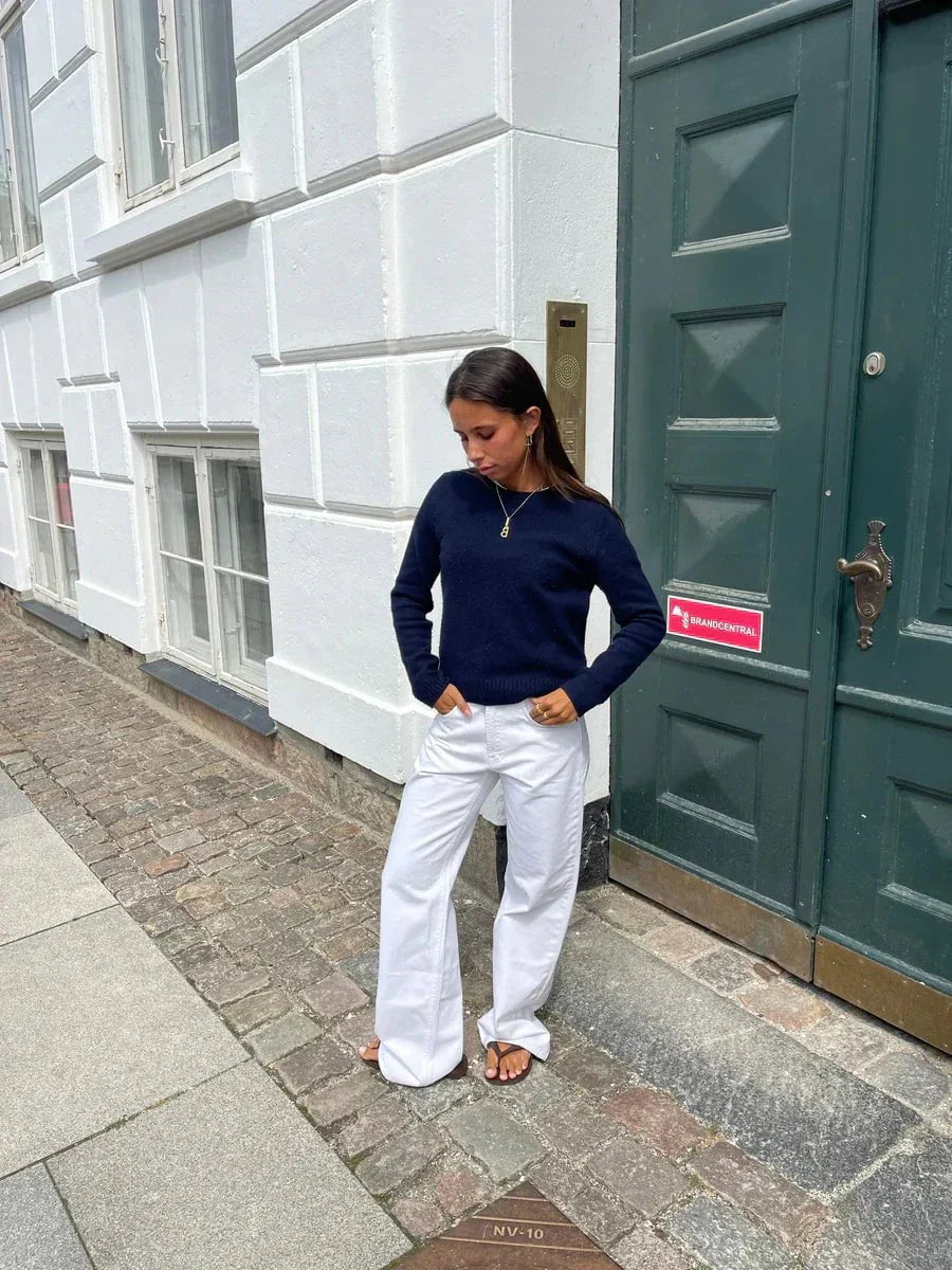 Jeune femme en pull bleu et pantalon blanc, posant contre une porte verte dans une rue pavée. Mode décontractée, style urbain, look automnal.