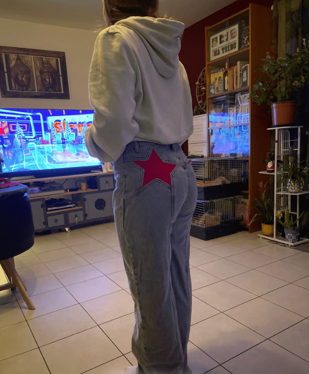 Jeune femme en jean avec étoile rouge à l'arrière, debout dans un salon moderne, regardant une émission télévisée sur un grand écran.