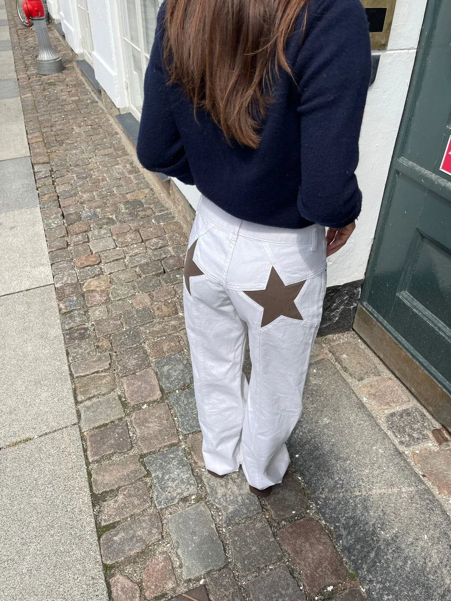 Femme portant un pantalon blanc avec des étoiles marron à l'arrière, marchant sur un trottoir pavé. Mode urbaine, style décontracté.