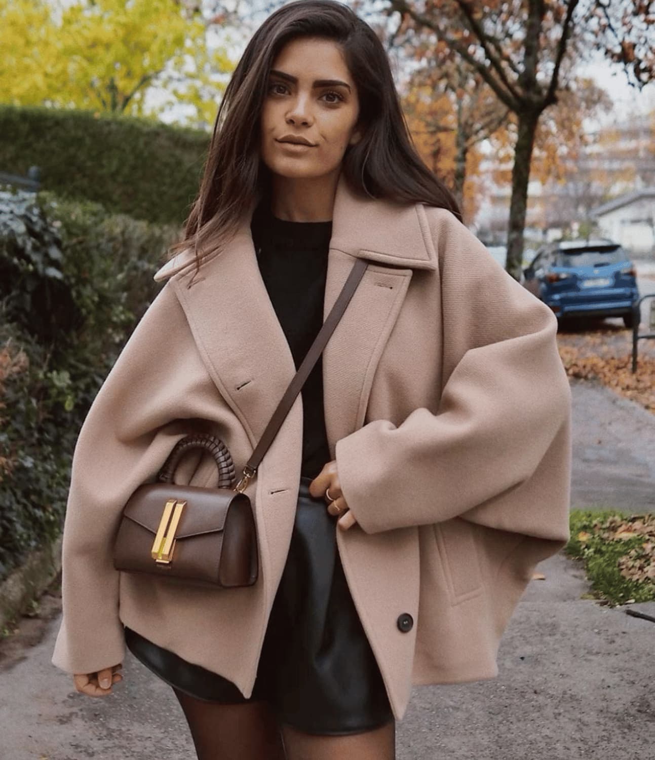 Femme portant un manteau beige oversize, sac à main marron, jupe noire, posant dans une rue automnale. Mode automne, style chic et élégant.