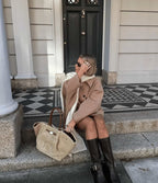 Femme assise sur des marches, portant un manteau beige et des bottes noires, avec un sac en laine beige. Devant une porte noire élégante.