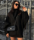 Femme élégante en manteau noir oversize, robe noire, lunettes de soleil, sac à main en cuir noir, posant devant une grille métallique décorative.