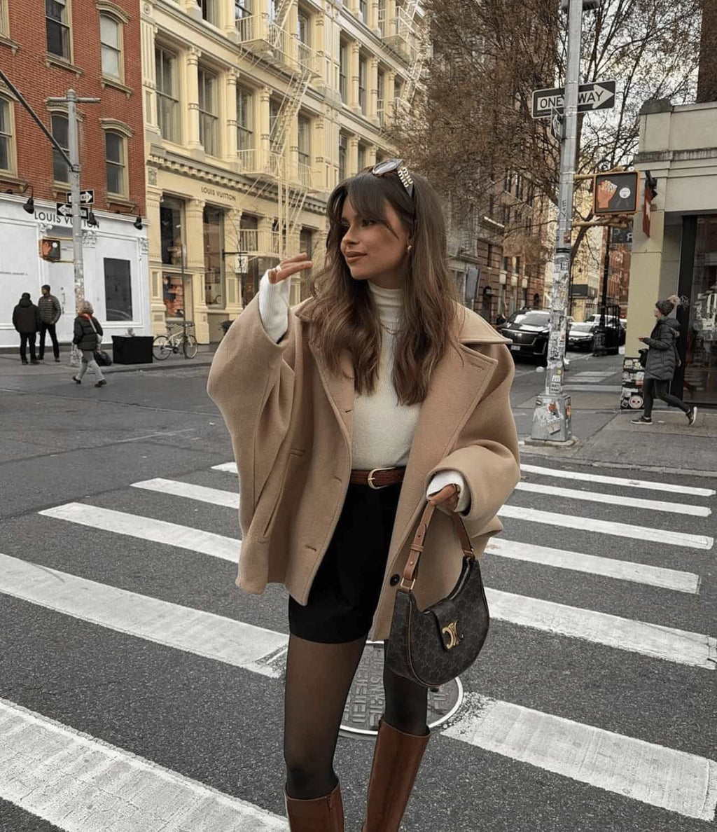 Jeune femme élégante traversant une rue urbaine, portant un manteau beige, un pull blanc, un short noir et des bottes marron, sac à main en cuir.