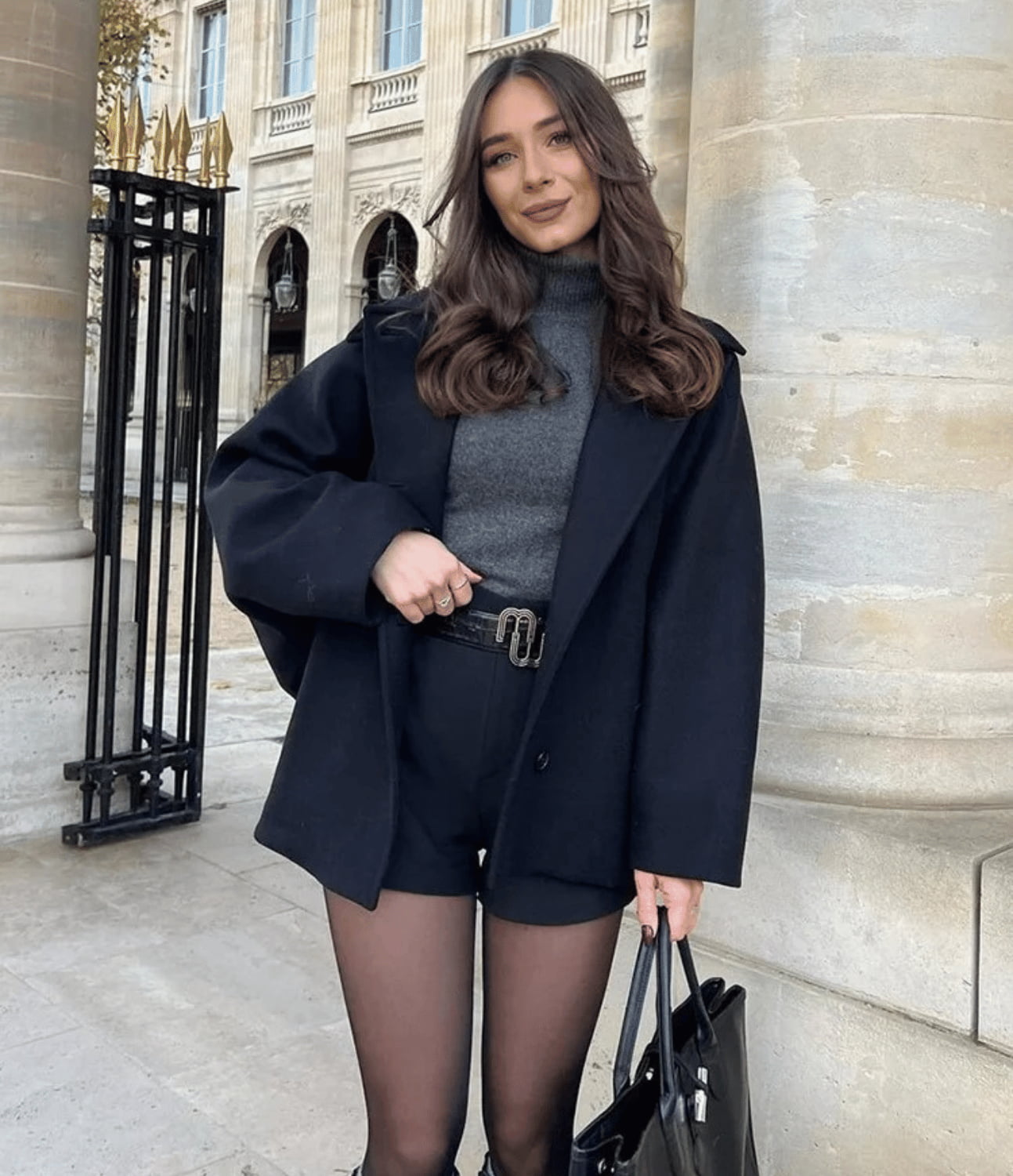 Jeune femme élégante en manteau noir, pull gris et short, posant devant un bâtiment historique à Paris, tenant un sac à main noir.