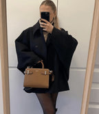 Femme en manteau noir oversize, tenant un sac à main marron élégant, se prenant en photo dans un miroir. Mode automne, style chic et moderne.