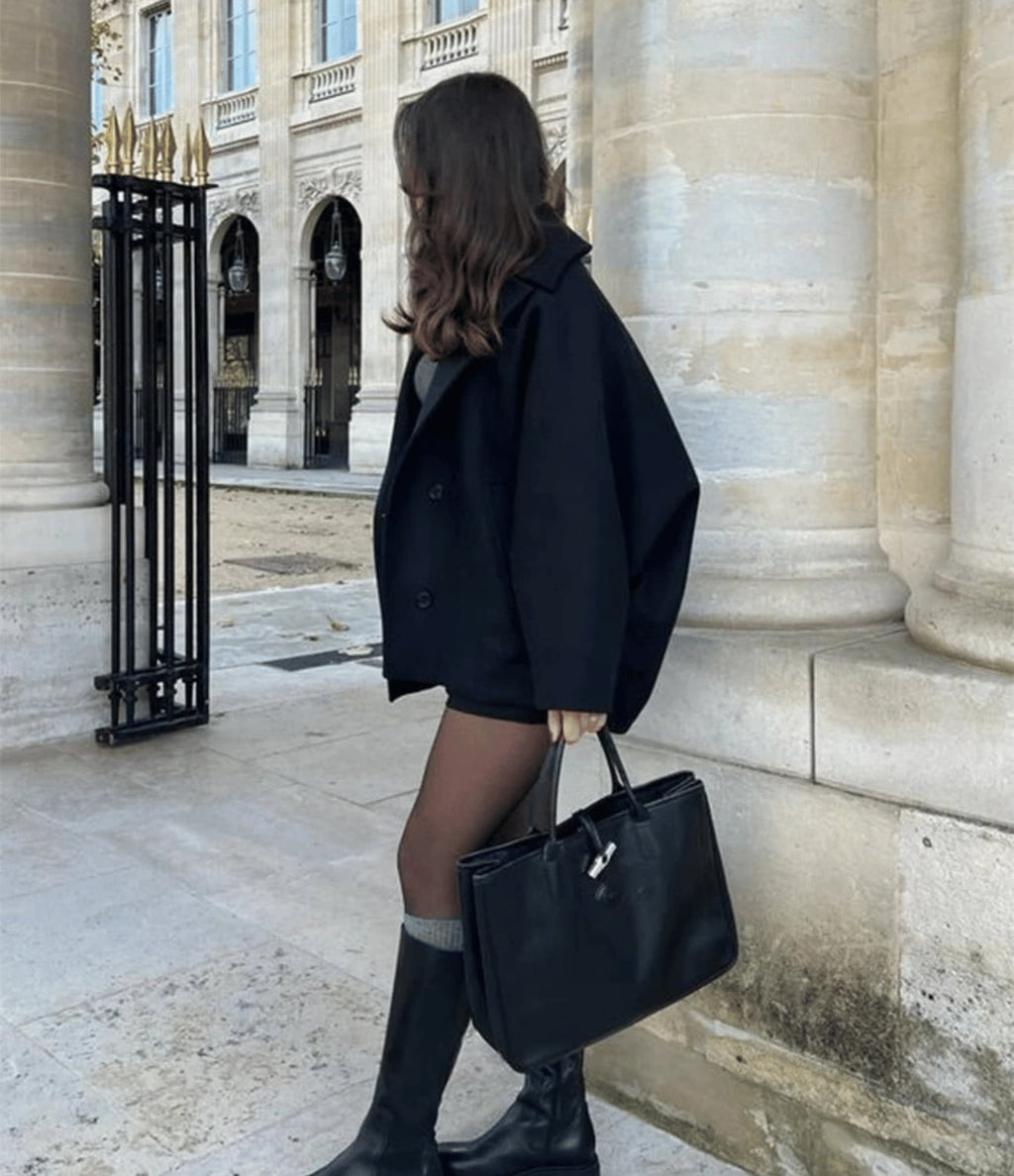 Femme élégante en manteau noir, jupe courte et bottes, portant un sac à main noir devant un bâtiment historique à colonnes. Mode automne chic.