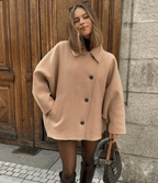 Femme portant un manteau beige oversize, posant devant une porte en bois. Mode automne, style chic, bottes noires, sac à main élégant.