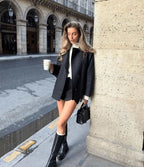 Jeune femme élégante en manteau noir, jupe courte et bottes, tenant un café à emporter, posant dans une rue parisienne historique.