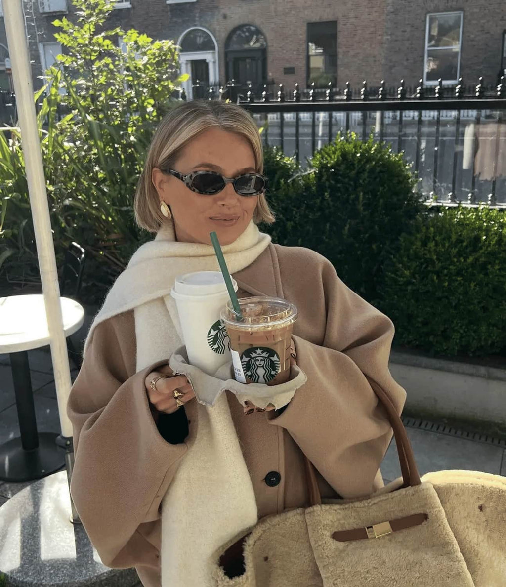 Femme élégante portant un manteau beige, lunettes de soleil, tenant des boissons Starbucks, sac en fourrure, devant un café en plein air.