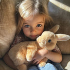 Petite fille aux yeux bleus tenant un lapin en peluche beige, assise sur un fauteuil beige. Enfant câlinant un jouet doux, ambiance chaleureuse.