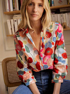 Femme portant une chemise à motifs floraux colorés, assise devant une étagère de livres. Mode féminine, style décontracté, tendance printemps.