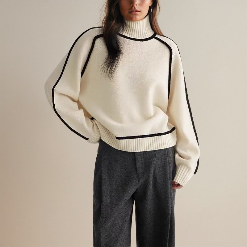 Femme portant un pull blanc à col roulé avec bordures noires, manches longues, associé à un pantalon gris. Mode automne-hiver élégante et moderne.