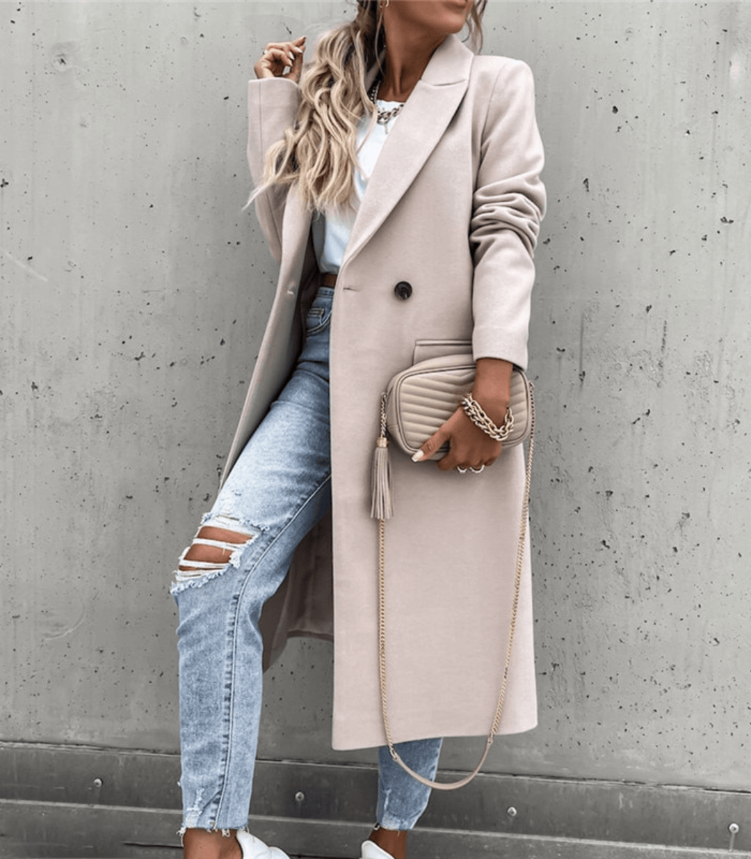 Femme en manteau beige élégant, jeans déchirés, sac à main beige, posant contre un mur gris. Mode automne, style urbain chic, tendance 2023.